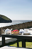 Erdbeerbiskuit und Teekanne auf einem Tisch mit Blick auf das Meer, Cornwall, UK