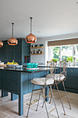Metal bar stools and brass pendant lights in teal fitted kitchen of Oxfordshire home UK