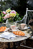 Geschnittene Pfingstrosen und Croissants mit Tee in Tassen auf einem Tisch im Garten in Surrey, Großbritannien