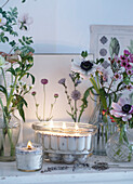 Verschiedene Glasvasen und Gläser mit Frühlingsblumen auf einem Regal mit botanischen Motiven und Kerzen