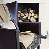 Brown leather armchair with throw and cushion in front of a fireplace stacked with wood