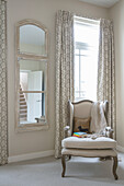 Light colored upholstered armchair in front of window with floor length curtains and wall mirror
