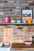 Wooden shelf with colourful bowls above kitchen worktop on brick wall