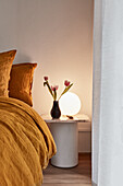 An apartment bedroom with turmeric crushed linen bedding, a marble side table, round lamp and a vase with pink tulips.