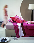 A magenta blanket and cushion on a bed with a plate of figs in the foreground