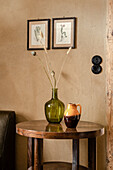 Green glass vase and a pitcher on a round side table in front of wall in earth tones