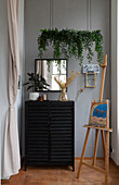 Easel, black chest of drawers and hanging plant in the hallway