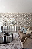 A velvet sofa with cushions, a silver chest of drawers with a round mirror above it in a wallpapered living room