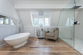A free-standing bathtub, a side table and an armchair with a shower behind a glass partition in a bright bathroom