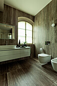 A spacious washstand, a toilet and a bidet in a bathroom with dark tiles