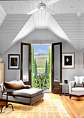 Chaiselongue, Sessel und Beistelltische im Zimmer mit weiß gestrichener Holzdecke und Balkon mit Landschaftsblick