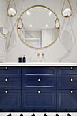 Blauer Waschtischunterschrank, darüber Marmorfliesen, runder Spiegel und Pendelleuchten im Bad