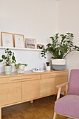 Sideboard mit Zimmerpflanzen, darüber Regal, im Vordergrund Sessel mit rosafarbenem Bezug