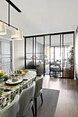 Long, oval dining table and upholstered chairs, in the background glass partition with sliding doors to living room