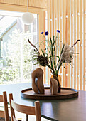 Sculptures and vase with a dried autumn bouquet on a round wooden tray on a dining table