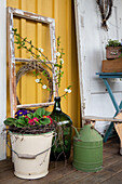 Emailleeimer mit Primeln (Primula), Ballonflasche mit Obstzweigen und Kranz aus Clematiszweigen an Hauswand