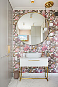 Pedestal washbasin in toilet with large round mirror and wall decorated with floral wallpaper