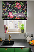 Black conglomerate counter top and sink, above window and roman shade with tropical pattern