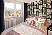 Bedroom with pink double bed, bedside table, and wallpaper with terrarium motif, window with black velvet curtains