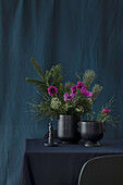Bouquet of various fir greenery with pink ranunculus in a black vase against a blue background