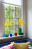 Green glass vase with forsythia branches and colored glass tea light holders on windowsill