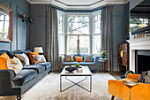 Klassisches Wohnzimmer mit blauen Wänden, farblich passendem Samtsofa, gelben Akzenten, Kamin und Erkerbereich
