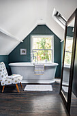 Freestanding bathtub and patterned armchair in attic bathroom with wooden floor