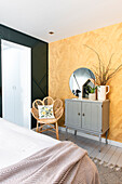 Bedroom with golden yellow plant wallpaper, rattan chair and grey chest of drawers