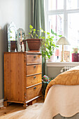 Wooden chest of drawers with plant and mirror in front of a bed