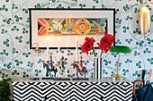 Kommode in Art-Deco-Stil mit Kerzen und Blumen, darüber Bild und gemusterte Tapete