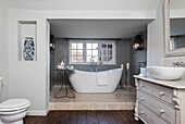 Freestanding bathtub in light grey bathroom with wooden floor and vintage chest of drawers