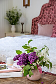 Fliederstrauß in rosafarbener Vase, Bett im Hintergrund