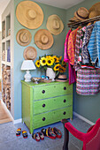 Hallway with green chest of drawers, straw hats on the wall and colourful clothes on the coat rack