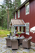 Terrassensitzgruppe mit Rattanmöbeln vor rotem Holzhaus im Wald