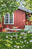 Gedeckter Gartentisch mit Blumenstrauß vor rotem Gartenhaus im Sommer