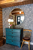 Round wall mirror above blue vintage chest of drawers, wooden chair next to it