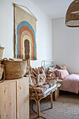 Wall hanging with rainbow motif, rattan furniture, wooden chest of drawers and bed in children's room