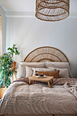 Bett mit Rattan-Kopfteil, Bettwäsche in Naturtönen, Frühstückstablett und Pflanze im hellen Schlafzimmer