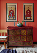 Antik bemalte Holzkommode mit asiatischem Flair vor orangefarbener Wand mit gerahmten Bildern