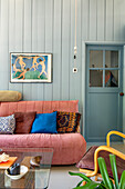 Wohnzimmer mit rosa Sofa und farbigen Kissen vor hellblauer Holzwand