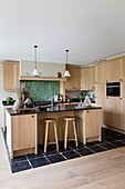 Kitchen with light-coloured wooden cabinets, green tiles, kitchen island and bar stools