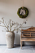 Wooden bench and decorative tree branch in vase with moss wreath on the wall