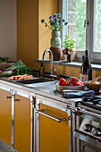 Kitchen with yellow cupboard doors and stainless steel surfaces, fresh vegetables and herbs on the worktop