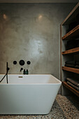 Free-standing bathtub in front of concrete wall and shelves with towels