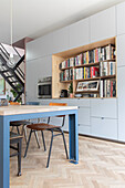 Open-plan kitchen with integrated bookshelf wall and dining table in blue and wood look