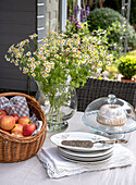 Laid garden table with apple basket, bouquet of daisies and cake