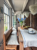 Rustic dining area with wooden table and bouquet of flowers, bench with linen cushions, glass bead chandelier