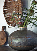 Steel vase with thistle flowers on a dark wooden board, rustic decoration in the background