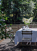 Tisch mit weißer Tischdecke und Blumen und Vintage Gartenstühle auf sonniger Terrasse