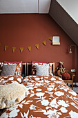 Sloping ceiling bedroom with patterned bed linen, rust-coloured wall and garland